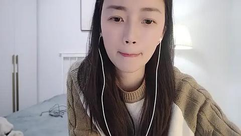 Media: Video of a young East Asian woman with straight black hair, wearing a beige sweater, seated indoors, with a white wall and lamp in the background.