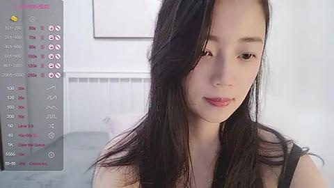 Media: A video of an East Asian woman with long, straight black hair and light skin, wearing a black top, smiling softly, in a brightly lit room with white walls.