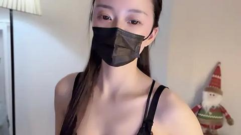 Media: Video of a young Asian woman with fair skin, long brown hair, wearing a black mask, black tank top, and a red and green Christmas elf decoration behind her.