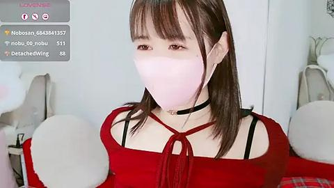 Media: Video of a young woman with pale skin, brown hair, and a red off-shoulder top, wearing a pink face mask. Background shows a blurred room with white furniture.