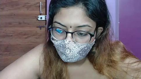 Media: Video of a South Asian woman with medium-brown skin, wearing glasses, a white patterned face mask, and a bindi. She has long, wavy, brown hair with lighter ends. Background shows a wooden door and a purple wall.