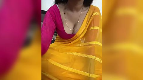 Media: Video of a woman with medium skin tone, wearing a vibrant orange saree with yellow borders, a pink blouse, and a gold necklace, taken indoors, blurred background.