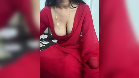 Media: Video of a woman with medium skin tone and long black hair, wearing a deep V-neck red saree, revealing her large breasts, with a black floral-patterned blouse underneath, sitting in a blurred background.