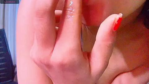 Media: A close-up video shows a fair-skinned woman's hand with red nail polish, fingers spread wide, obscuring her face. The background is blurred, featuring a dark-colored object and white curtains.