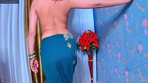 Media: Video of a topless woman with fair skin, wearing a teal sari with embroidered floral patterns, adorned with red and green bangles, standing in a room with blue floral wallpaper and a red rose bouquet.