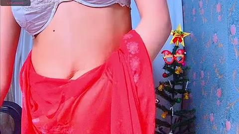 Media: Video of a light-skinned woman with a small mole on her abdomen, wearing a white bra and a red sheer sarong. Background shows a Christmas tree with festive decorations and a blue wall.