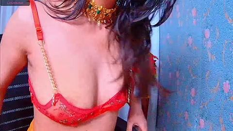 Media: Video of a woman with medium brown skin and long, wavy dark hair wearing a red lace bra with gold chain detailing, a gold necklace, and a blue floral-patterned wall in the background.