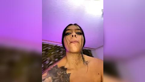 Media: A video of a topless woman with dark hair and tattoos, standing in a dimly lit room with purple lighting. She has a serious expression and appears to be wearing dark makeup.