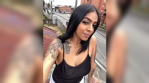 Media: Video of a young Latina woman with long black hair and tattoos, wearing a black tank top, taking a selfie in an urban street setting.