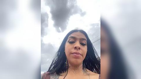 Media: A video of a Latina woman with long black hair, medium skin tone, wearing a beige top, standing in a cloudy sky.