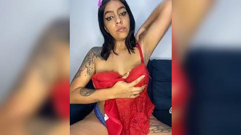 Media: A video of a Latina woman with medium brown skin and black hair, wearing a red lace bra and blue panties, sitting on a couch. She has tattoos on her left arm.