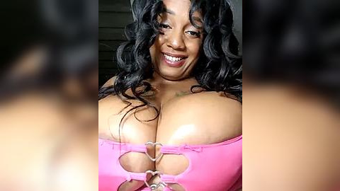 Media: Video of a smiling, curvy Black woman with large breasts, wearing a pink, heart-shaped bikini top with a silver ring. Her wavy black hair frames her face. Background is blurred.