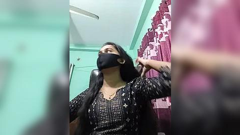 Media: A video shows a woman with long black hair, wearing a black face mask, a black patterned dress, and a black headscarf, seated in a room with teal walls and pink floral curtains.