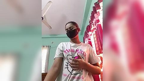 Media: Video of a woman in a white t-shirt with a Hello Kitty design, wearing a black face mask, standing in a room with teal walls and red curtains.