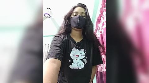 Media: A video of a smiling woman with long black hair, wearing a black face mask, black T-shirt with a cartoon cat graphic, and standing in a brightly lit bathroom with pink towels and a white showerhead.