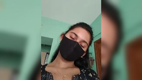 Media: Video of a young woman with light brown skin, dark hair, and a black mask, wearing a black floral-patterned dress, indoors with light green walls and a wooden door.