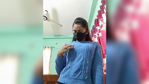 Media: A video of a young woman taking a mirror selfie in a bathroom with mint green walls, wearing a blue knitted sweater, black face mask, and long dark hair pulled back.