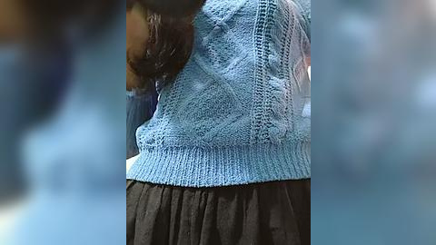 Media: Video of a woman's torso, wearing a blue knitted sweater over a black skirt, with blurred background.