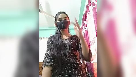 Media: A video of a woman with long black hair wearing a black face mask, black dress with white patterns, and holding her hair up, taken in a room with teal walls and pink curtains.