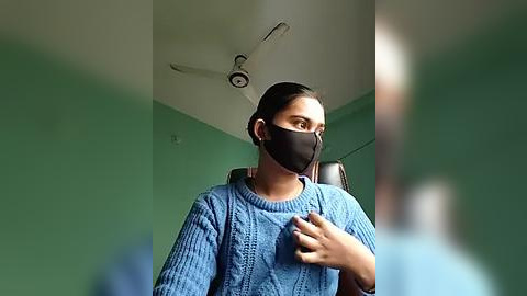 Media: Video of a woman in a blue sweater with a black face mask, standing in a room with green walls and a ceiling fan.