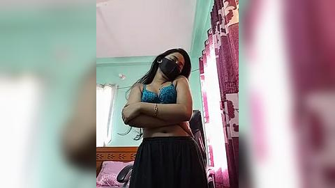 Media: Video of a woman with long black hair, wearing a blue lace bra, black mask, and loose black pants, standing in a bright, mint-green bedroom with pink curtains and a bed.