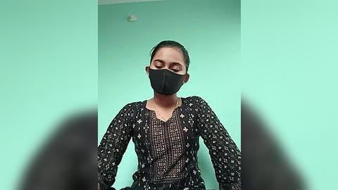 Media: A video of a South Asian woman with light brown skin, wearing a black face mask, black blouse with white floral patterns, and a green wall background.