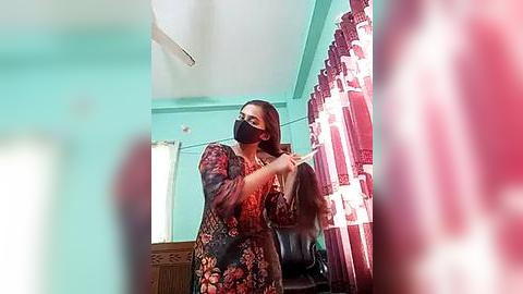 Media: Video of a woman in a floral dress, black mask, and glasses, adjusting her hair in front of a red-patterned curtain, teal-painted wall, and wooden furniture.