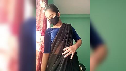 Media: Video of a young woman with medium-dark skin tone, wearing a blue blouse, black saree, and black mask, standing in a room with green walls and a red curtain.