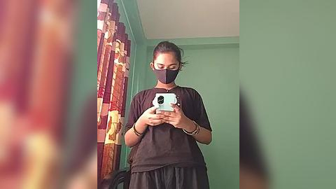 Media: Video of an Asian woman with dark hair in a ponytail, wearing a black face mask and a dark t-shirt, holding a phone, against a green wall with a patterned curtain.