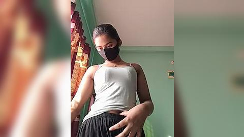 Media: Video of a young, fit woman with medium skin tone, wearing a gray tank top and black mask, standing in a room with teal walls, red curtains, and a green chair.