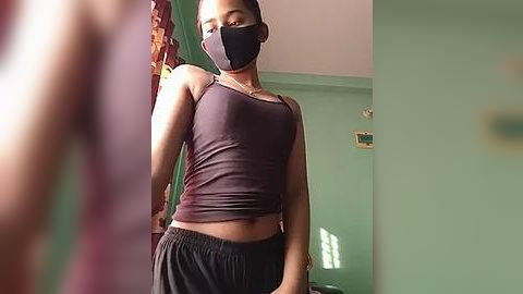 Media: A video of a slender Black woman with a medium complexion wearing a black face mask, dark tank top, and loose black pants. She stands in a dimly lit, mint-green room with a door and a window partially visible.