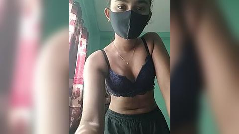 Media: Video of a young woman with medium skin tone, wearing a black mask, lace bra, and loose pants, standing in a dimly lit room with teal walls and a red-patterned curtain.