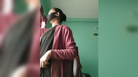 Media: Video of a woman in a mask, maroon cardigan, and black shirt, standing in a green-painted room with a white ceiling and a smoke detector.