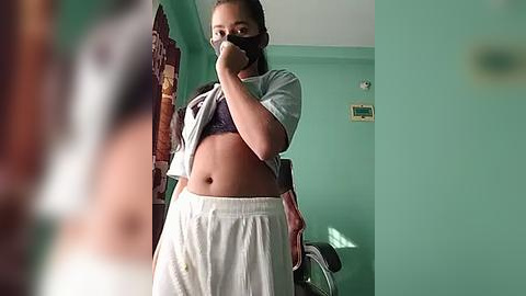 Media: A video of a young woman in a teal bathroom, lifting her shirt to reveal a purple bra, wearing white shorts. She holds a mask in her hand, capturing a mirror reflection.