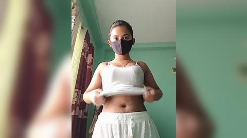 Media: Video of a young woman with medium skin tone, wearing a black face mask and white tank top and shorts, standing in a teal-painted room with green curtains.