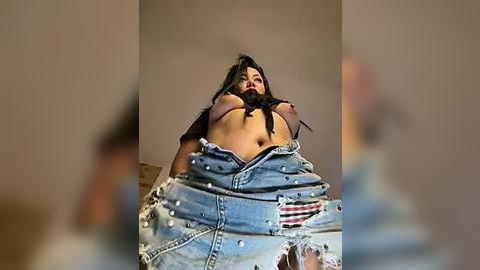 Media: Video of a woman with long black hair, wearing torn denim shorts and a black top, exposing her breasts. She appears distressed, in a dimly lit room with blurred background.