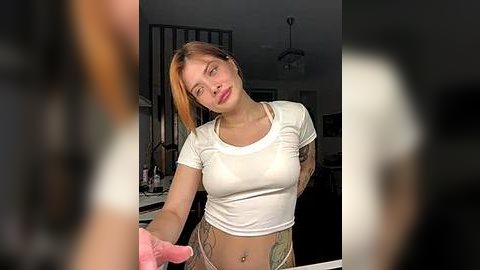 Media: A video of a young, fair-skinned woman with short, dyed orange hair, wearing a white crop top and patterned pants, standing in a dimly lit kitchen with a blurred figure in the background.