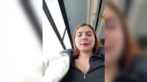 Media: Video of a young woman with fair skin, medium-length red hair, and light makeup, wearing a dark jacket, relaxing on a bus seat with a white cushion, eyes closed, serene expression.