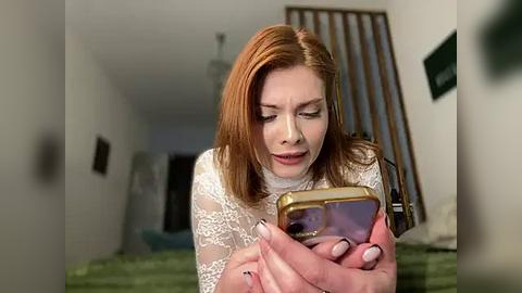 Media: Video of a red-haired woman in a white lace top, holding a phone, in a dimly lit hallway with green walls and a bed in the background.