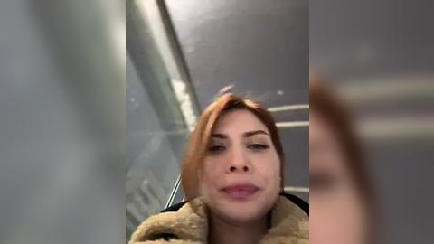 Media: Video of a fair-skinned woman with red hair, wearing a beige coat, making a silly face while looking at herself in a reflective surface, blurred background.