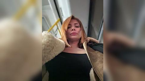 Media: Video of a blonde woman in a black top and tan jacket, leaning against a bus window, with blurred yellow pole and bus seat in the foreground.