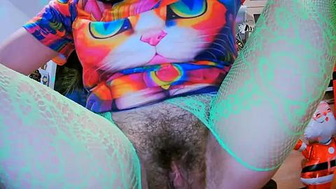 Media: Video of a person wearing a vibrant, psychedelic shirt featuring a colorful cat face, green fishnet stockings, and visible pubic hair, seated with legs spread, capturing a playful, provocative pose.