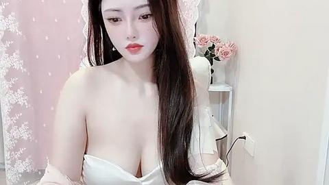 Media: Video of an East Asian woman with long, straight black hair, fair skin, and full lips. She wears a strapless white dress, sitting in a room with pastel pink curtains and a white shelf with flowers.