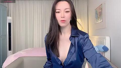 Media: A video of a young Asian woman with fair skin and long black hair, wearing a partially unbuttoned blue satin shirt, posing in a modern bedroom with beige curtains, a white bed, and a wall art.