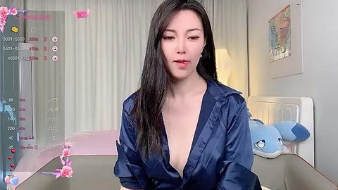 Media: Video of an Asian woman with long black hair, wearing a blue satin robe, sitting at a table in a pastel-colored bedroom.