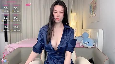 Media: Video of an East Asian woman with long black hair, fair skin, wearing a dark blue satin blouse exposing cleavage, sitting on a bed in a modern bedroom with white walls and plush toys.