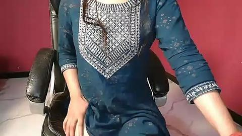 Media: Video of a woman seated on a black office chair, wearing a dark blue kurta with intricate white embroidery on the chest, against a red wall, with a light gray floor.