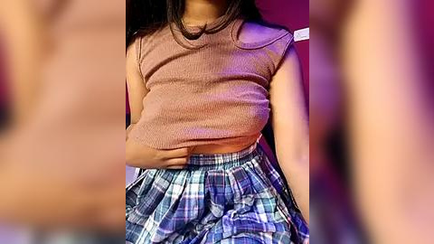 Media: Video of a young woman with light brown skin, wearing a cropped, ribbed, beige top and a high-waisted, blue plaid skirt, seated with her left hand on her abdomen. Background is blurred, featuring warm lighting.