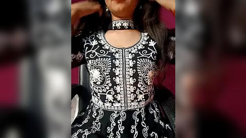 Media: Video of a woman with medium-brown skin and long, wavy dark hair, wearing a black and white embroidered dress with a high collar, seated on a black chair against a blurred maroon background.