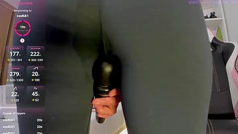 Media: Video of a person in a dark green bodysuit with a black dildo inserted anally, holding a bottle of wine. Background includes a partially visible chair and a wall with shelves.